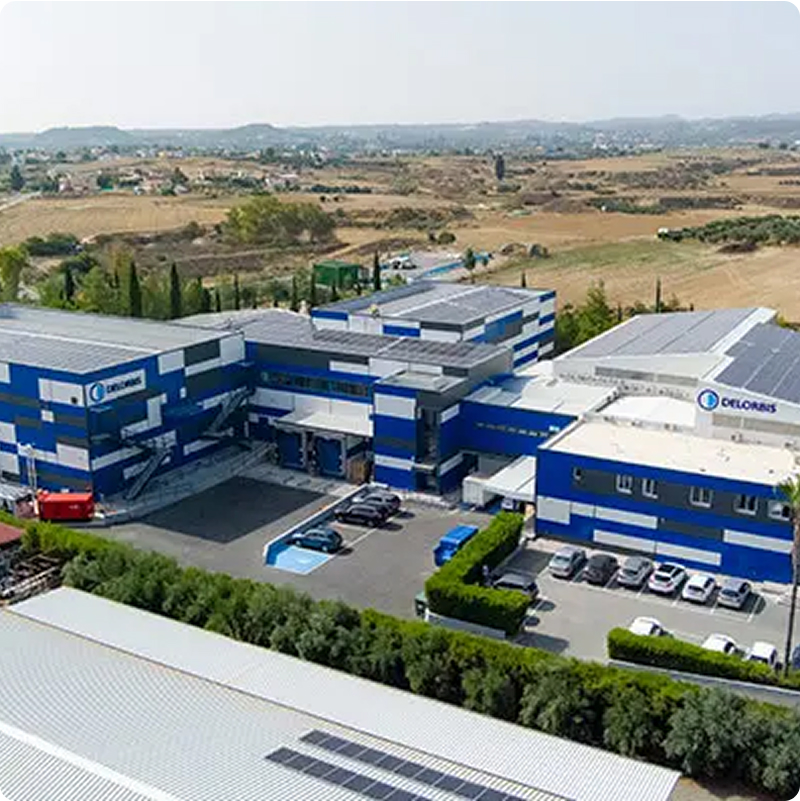 Tiefenbacher Group - Aerial view of a large blue and white industrial building with DELORIS logos, surrounded by parked cars and fields, highlighting its role in pharmaceutical healthcare solutions within a rural landscape.