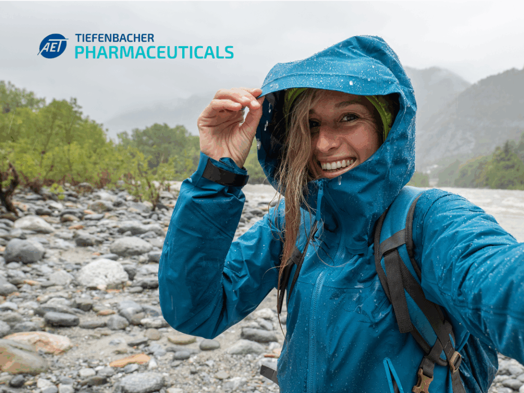 Tiefenbacher Group - A smiling woman in a blue rain jacket takes a selfie outdoors by a rocky river, holding her hood with one hand. Trees and misty mountains are in the background. The Tiefenbacher Pharmaceuticals logo highlights patient-centric drug development.