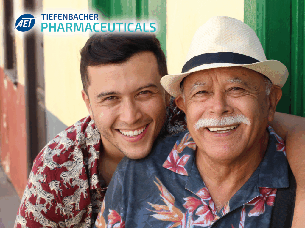 Tiefenbacher Group - A young man and an older man in patterned shirts and a hat smile together outdoors. The AET Tiefenbacher Pharmaceuticals logo appears in the top left corner, highlighting expertise in pharmaceutical healthcare solutions.