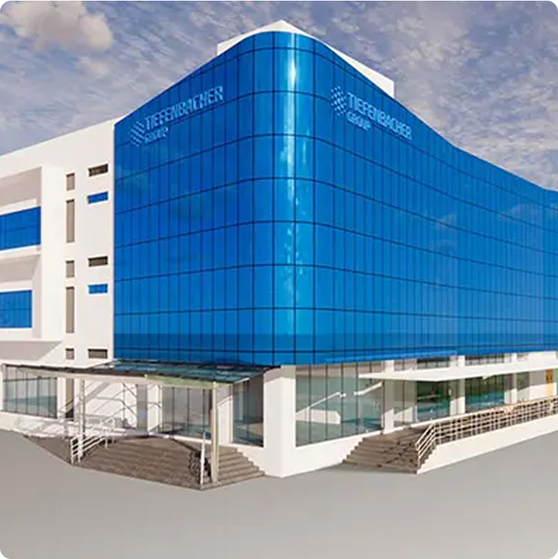 Tiefenbacher Group - Modern white building with a large blue-glass facade, home to TIEFENBACHER, a family-owned pharmaceutical company focused on patient-centric drug development. The entrance features stairs and a glass canopy under a partly cloudy sky.