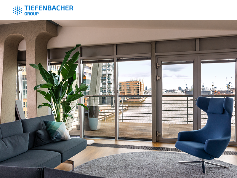 Tiefenbacher Group - Modern office lounge with blue and gray seating, a large plant, and floor-to-ceiling windows overlooking a river and cityscape; Tiefenbacher Group—a family-owned pharmaceutical company focused on patient-centric drug development—logo at the top.