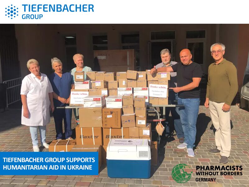 Tiefenbacher Group - Six people stand behind a large stack of medical supply boxes outside a building. Logos for Tiefenbacher Group and Pharmacists Without Borders Germany highlight pharmaceutical healthcare solutions supporting humanitarian aid in Ukraine.
