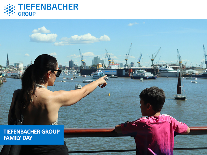 Tiefenbacher Group - A woman points towards the harbor while a young boy stands beside her, both gazing at the water with cranes, ships, and city buildings in the background—reflecting a commitment to patient-centric drug development. Image branded with "TIEFENBACHER GROUP FAMILY DAY.