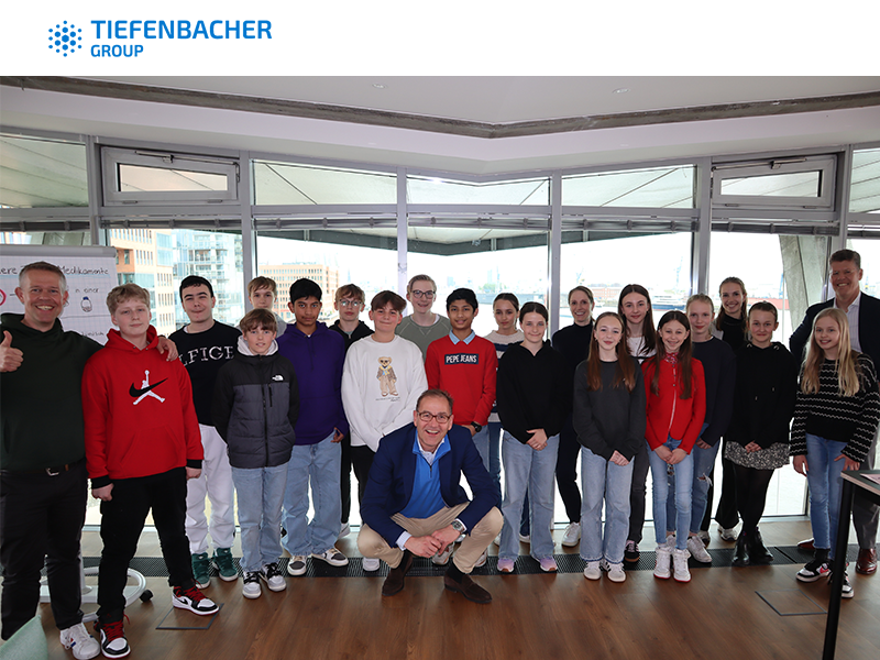 Tiefenbacher Group - A group of teenagers and adults pose together indoors in front of large windows. One adult kneels in front, another gives a thumbs up, and the Tiefenbacher Group logo—a family-owned pharmaceutical company focused on patient-centric drug development—is visible above.