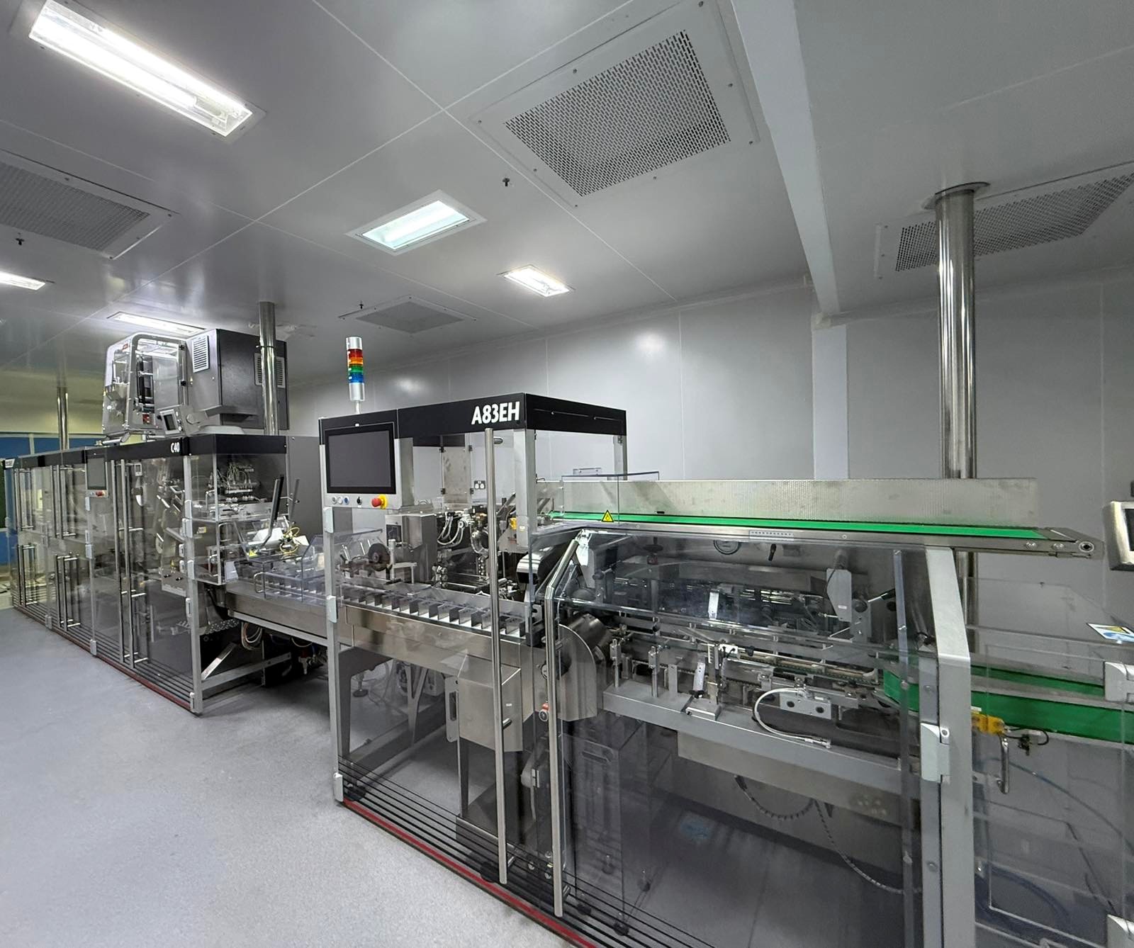 Tiefenbacher Group - A modern packaging machine for pharmaceutical healthcare solutions operates in a clean, well-lit factory with gray floors, white ceilings, transparent panels, conveyor belts, and colored indicator lights.