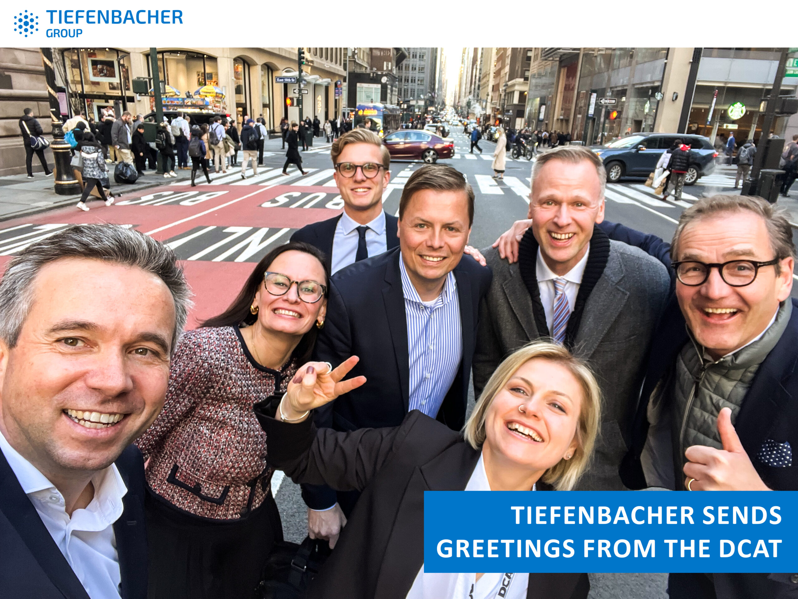 Tiefenbacher Group - A group of seven smiling people in business attire take a selfie on a busy city street. A banner reads, "Tiefenbacher sends greetings from the DCAT." The TIEFENBACHER GROUP logo highlights this family-owned pharmaceutical company.