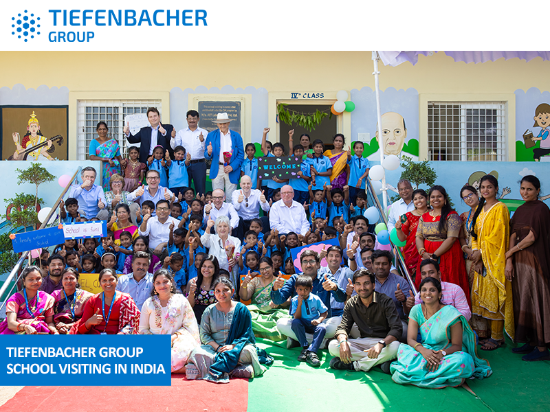 Tiefenbacher Group - A large group of children and adults, including visitors from the family-owned pharmaceutical company Tiefenbacher Group, pose together outside a decorated school building in India. The festive scene features balloons, welcome signs, and colorful attire.