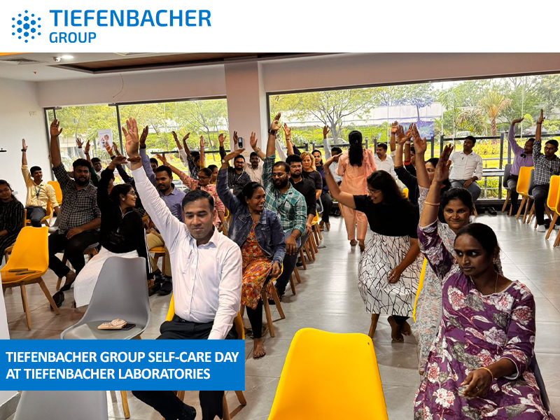 Tiefenbacher Group - A group of people, seated on yellow chairs indoors, raise their hands and smile during a self-care day event at Tiefenbacher Laboratories, a family-owned pharmaceutical company. Large windows show greenery outside.