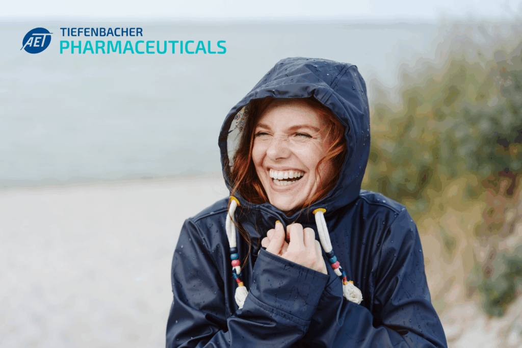 Tiefenbacher Group - A smiling woman in a navy raincoat stands on a beach, holding her hood. The AET TIEFENBACHER PHARMACEUTICALS logo appears in the top left, reflecting their focus on patient-centric drug development. The background shows the sea and greenery.