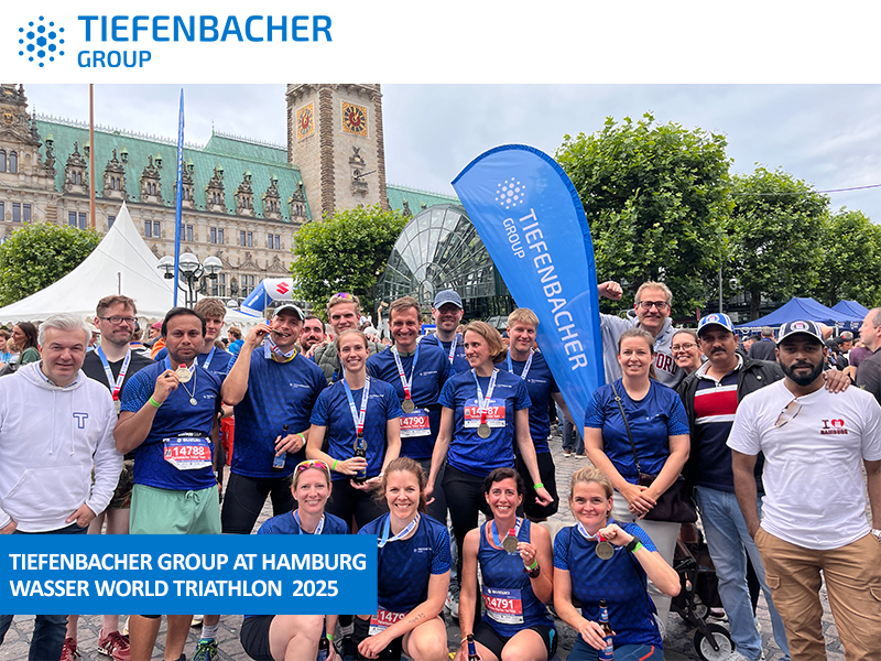 Tiefenbacher Group - A group of athletes in blue Tiefenbacher shirts, representing a family-owned pharmaceutical company, pose together, smiling and holding medals after the Hamburg Wasser World Triathlon 2025, with a crowd, banners, and historic buildings in the background.