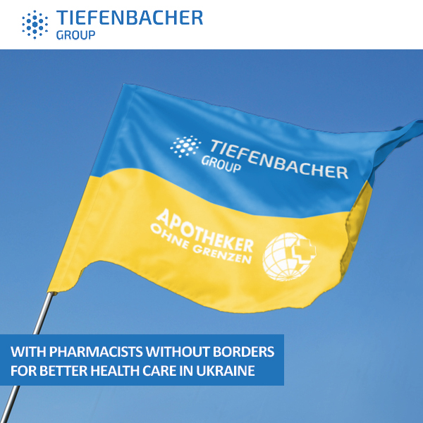 Tiefenbacher Group - A blue and yellow flag featuring the Tiefenbacher Group and Apotheker Ohne Grenzen logos waves against the sky. Text below reads: “With Pharmacists Without Borders for better pharmaceutical healthcare solutions in Ukraine.”.