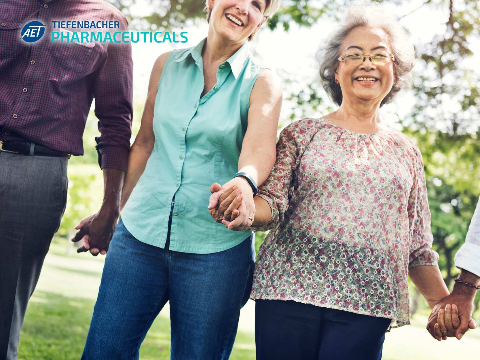 Tiefenbacher Group - Four adults stand outdoors holding hands and smiling in a bright, cheerful setting with green trees. The "Tiefenbacher Pharmaceuticals" logo appears in the upper left, highlighting expertise in pharmaceutical healthcare solutions.