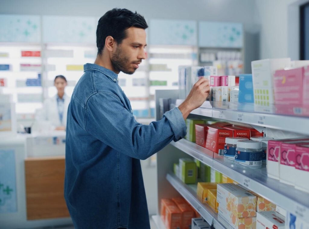 Tiefenbacher Group - A man in a denim shirt browses medicine and health products on a shelf in a pharmacy, while a pharmacist stands in the background behind the counter, highlighting access to finished dosage forms and API distribution.